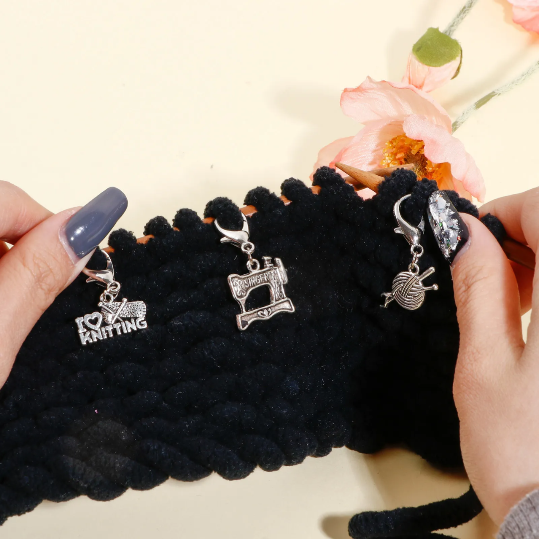 In hands, a black knit fabric is decorated with three silver knitting-themed charms from the "Stitch Markers 12pcs" set: "I love knitting," a sewing machine, and a ball of yarn with needles. These charms are crafted for precision stitch marking and feature durable steel construction, making them ideal for passionate crafters.