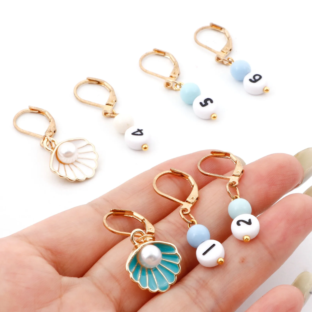 In the 29-Piece Crocheting Kit: Perfect for Beginners, you'll find a complete set of golden stitch markers. Each marker is embellished with beads numbered 1-6, and two of them showcase enchanting shell designs adorned with pearls.