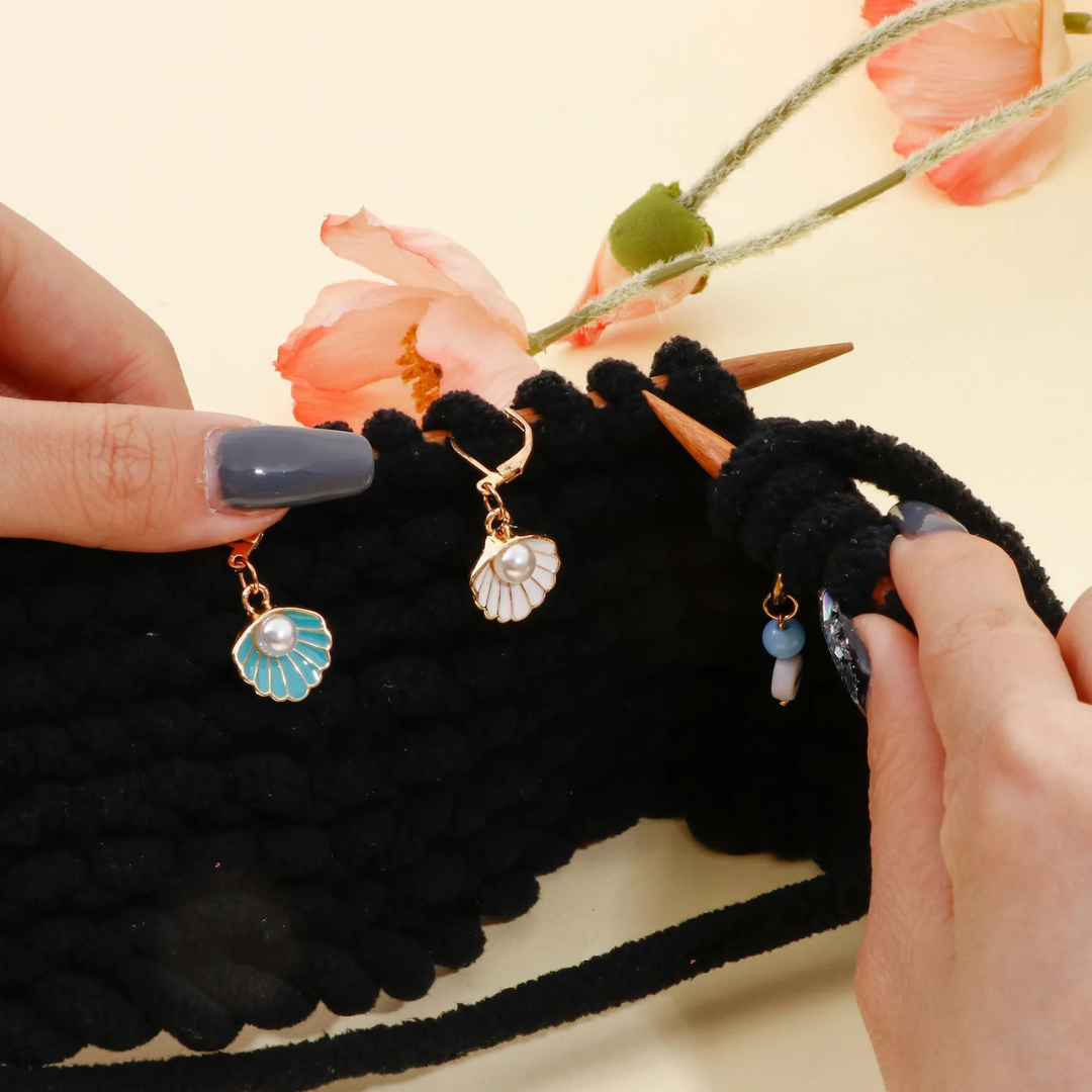 Hands knitting with chunky black yarn, embellished with Stitch Markers 9pcs, featuring shell pendants and orange flowers in the background, creating an elegant design that captivates the eye.