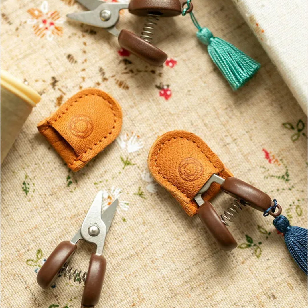 Small Scissors featuring brown handles and leather sheaths are elegantly displayed on a floral-patterned fabric surface next to a blue tassel, highlighting their precision and compact design.