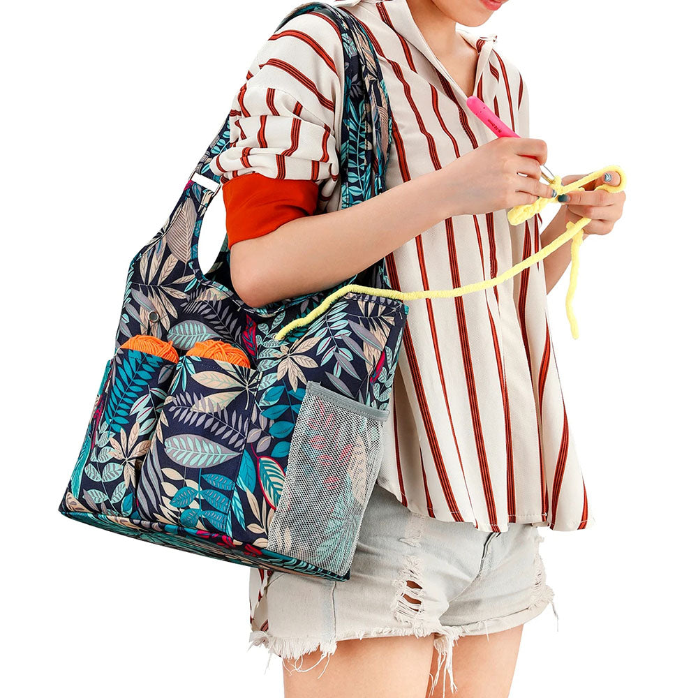 A person in a striped shirt and shorts holds yellow yarn while carrying a knitting accessory—a "Knit on The Go" Yarn Tote Bag (bundle) with partially visible yarn in the pockets, showcasing its blue floral pattern.