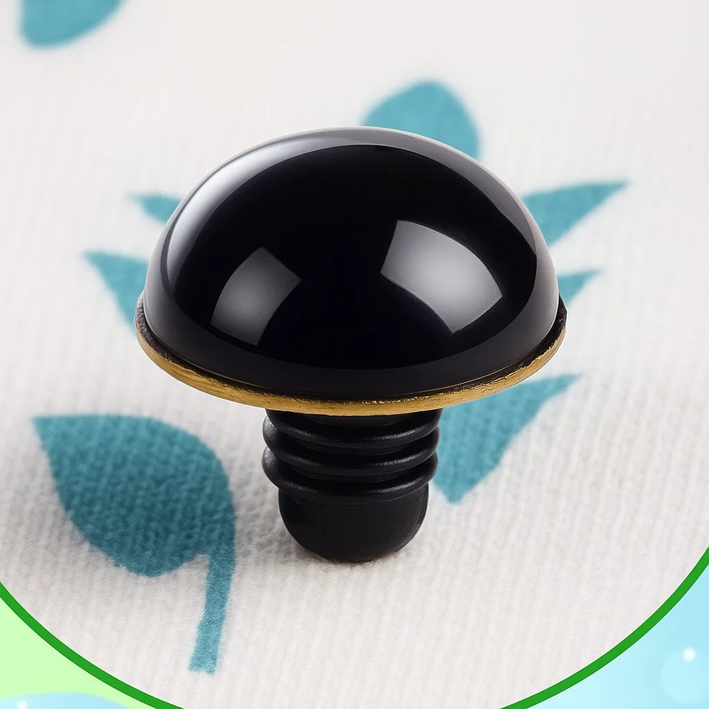A glossy black plastic eye with a gold backing from the Safety Eyes & Noses Set, ideal for crochet or amigurumi projects, displayed on white fabric with blue leaf patterns.