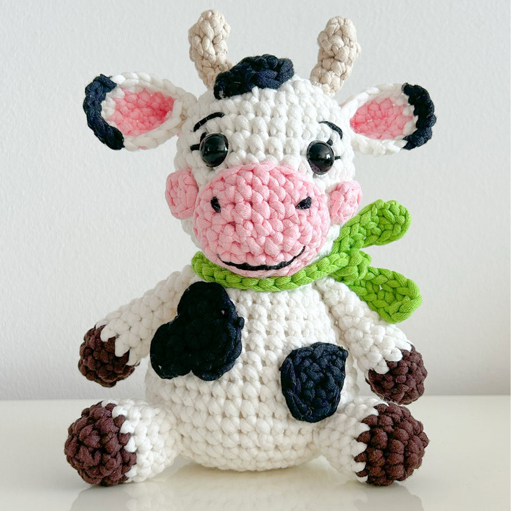 Hazel The Cow Crochet Pattern is an advanced beginner project featuring a crocheted cow with black spots, pink nose, big black eyes, brown hooves, beige horns, and a green scarf, shown sitting upright against a plain background.