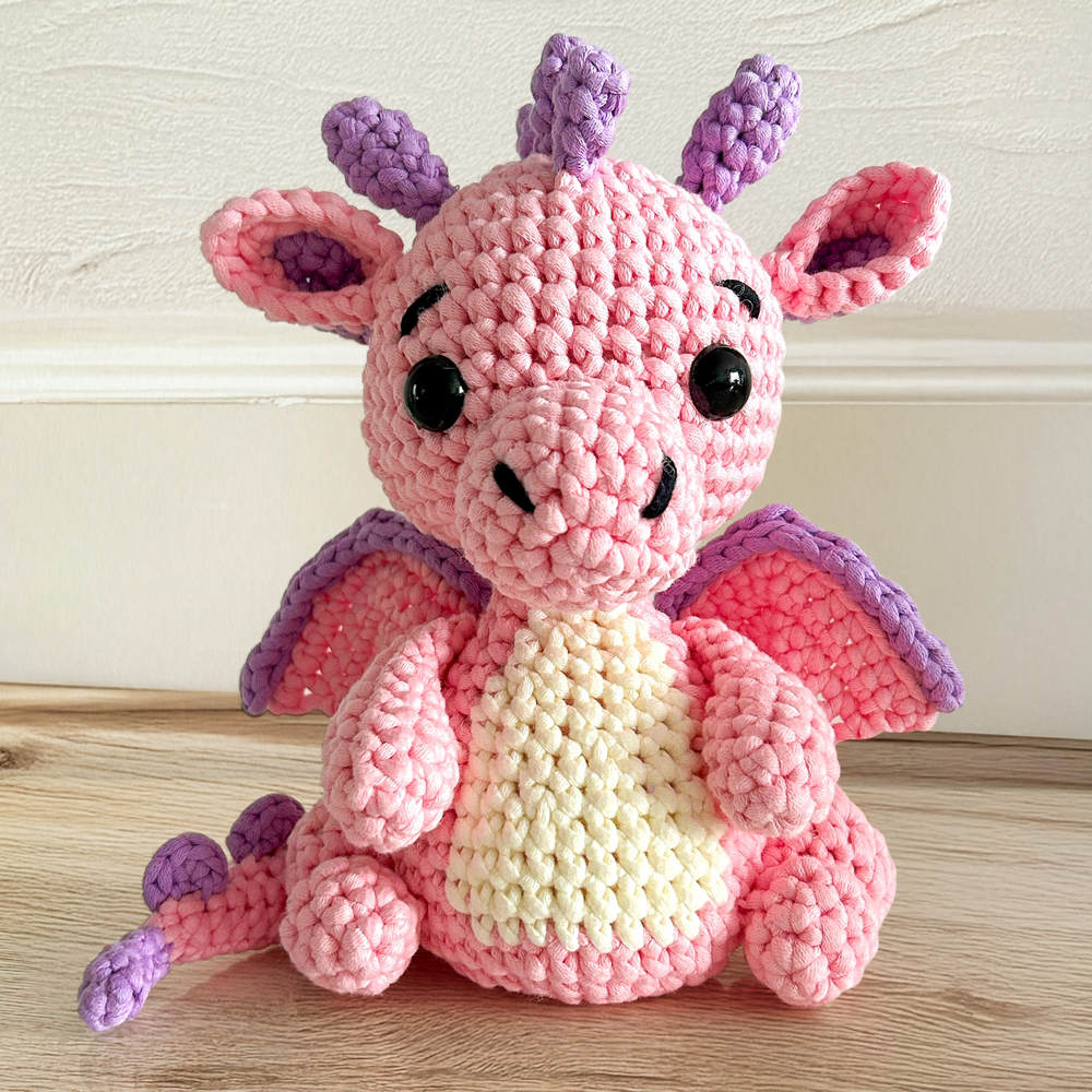Sparkle The Dragon Crochet Pattern features a pink and purple dragon plush toy with wings, shown sitting on a wooden surface against a white background.