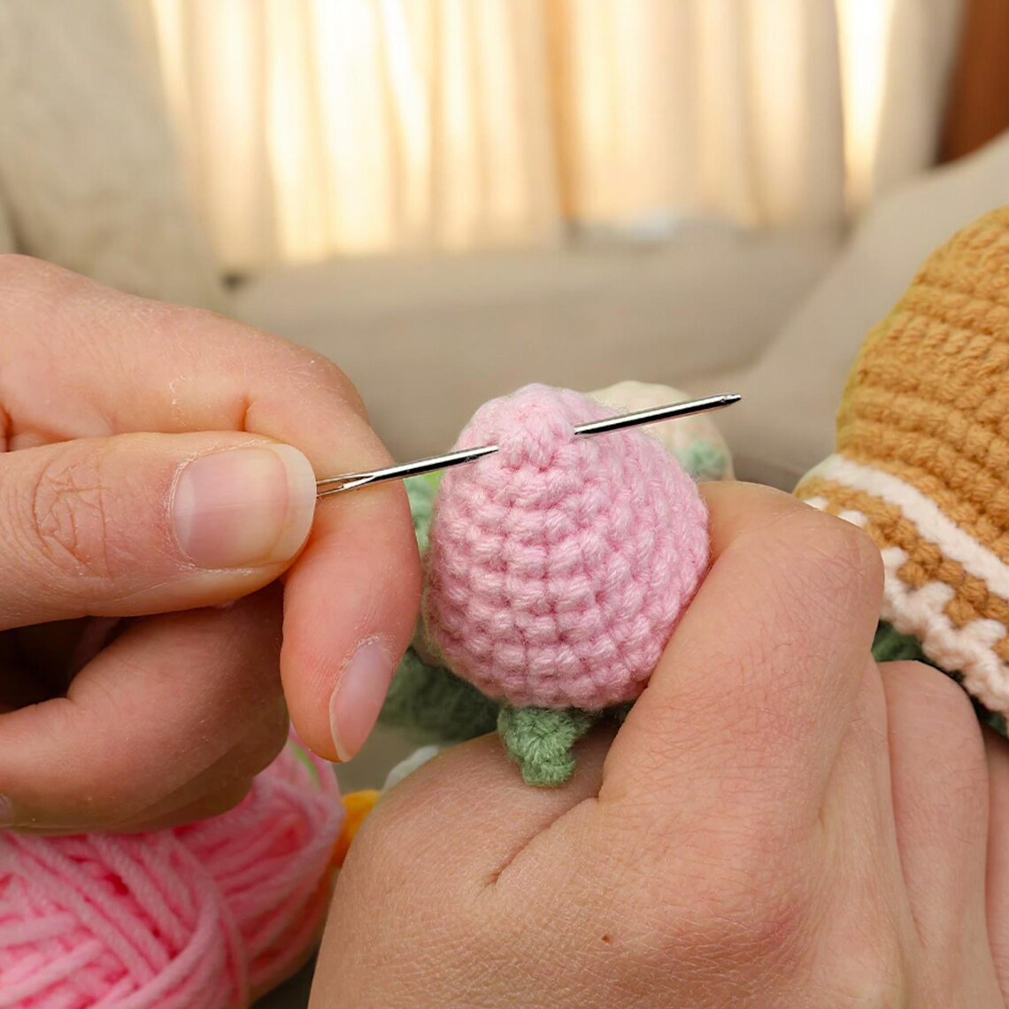 A person uses a needle to sew pieces of pink crocheted yarn from the Crochet Strawberry Plant Kit, with other crochet projects and yarn visible in the background.