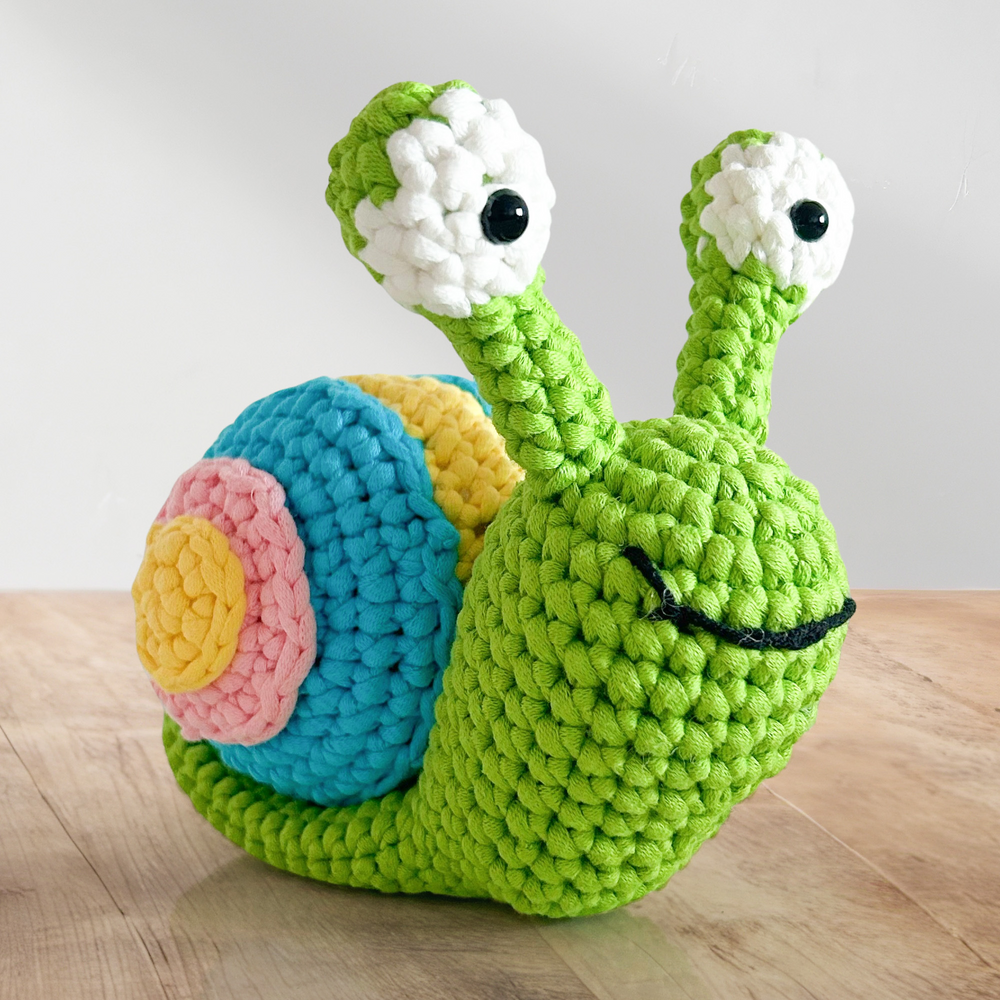Speedy The Snail Crochet Pattern features a green-bodied snail with big white eyes and a colorful shell in blue, pink, and yellow segments—ideal for anyone looking to crochet an adorable snail.