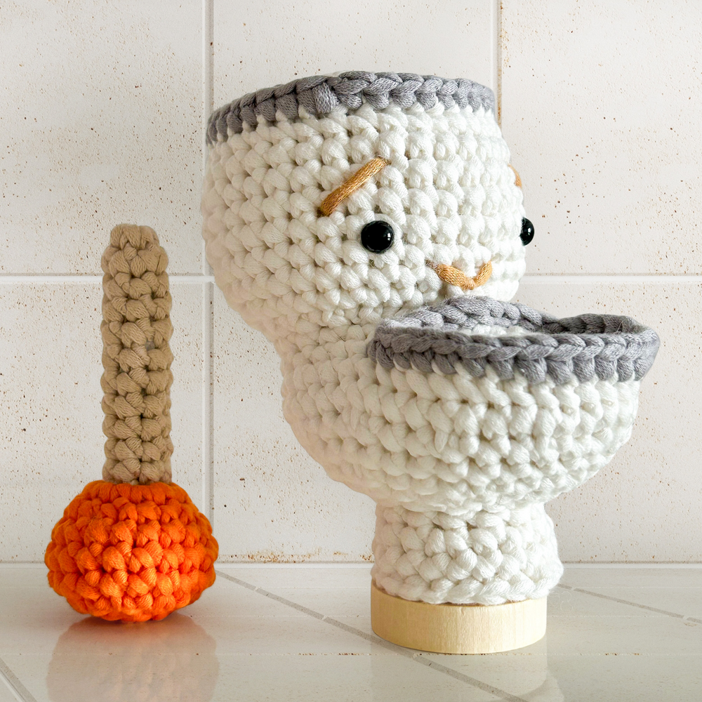 Splashy The Toilet Crochet Pattern creates a funny crocheted toilet with a face, shown beside a plunger with an orange base on tiles—a whimsical scene made from this playful pattern.