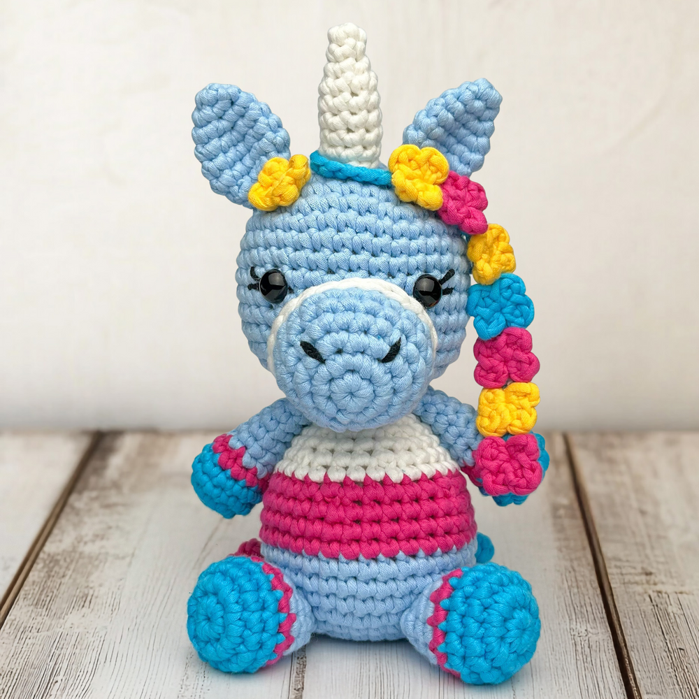 Twinkle the Unicorn Crochet Pattern creates a cute plush toy with a blue, white, and pink body, yellow and pink details, and a single horn. The unicorn is shown sitting on a wooden surface with a light background.
