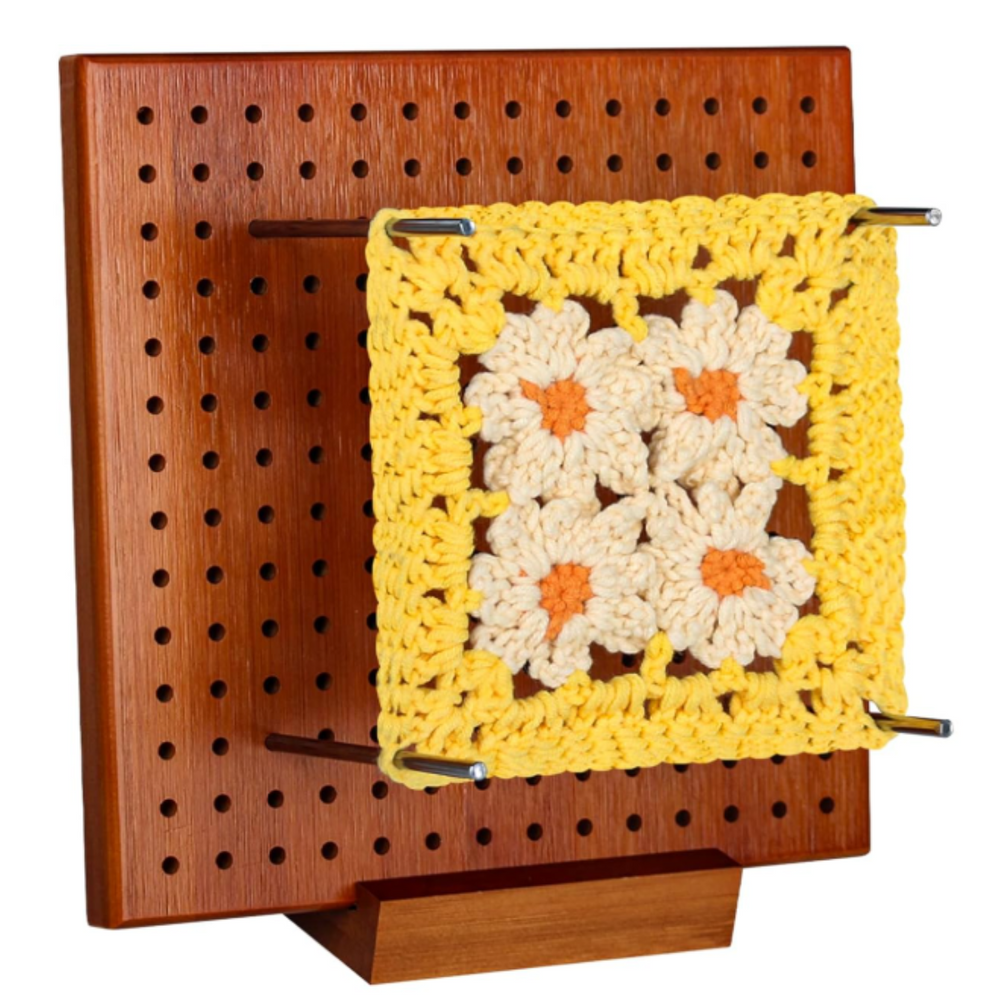 A Crochet Blocking Board holds a yellow and white flower granny square in place with metal rods, offering perfect shaping. The board features sturdy rubber wood construction for reliable use.
