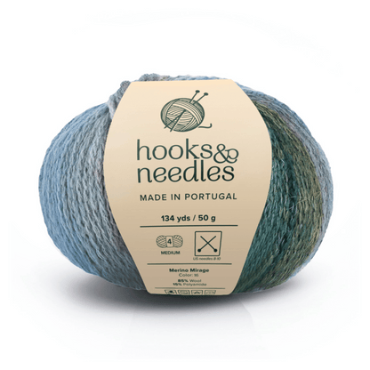 A 50g, 134-yard ball of Merino Mirage luxury yarn (85% Merino wool, polyamide) in blue-green variegated "Mirage" color, labeled hooks & needles, made in Portugal.
