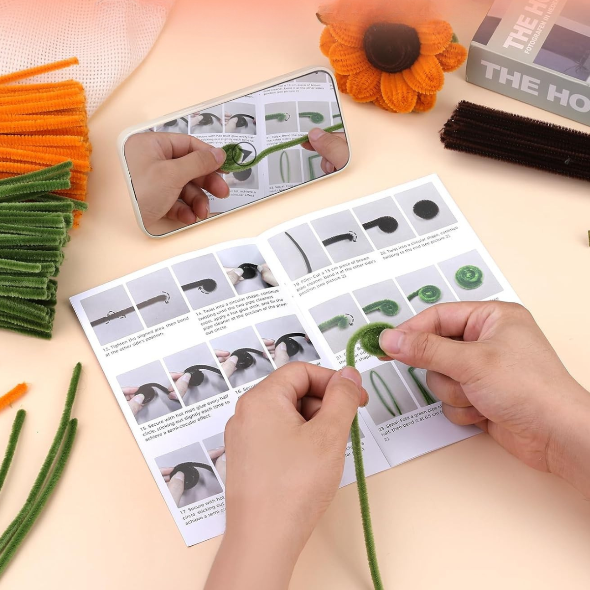 Hands shape a green pipe cleaner while following illustrated instructions in the Sunflower Crochet Kit booklet; craft supplies, a mirror, and a flower decoration are on the table.