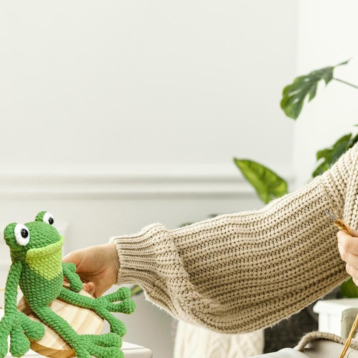 A person in a beige knitted sweater holds an adorable frog made with the Crochet Frog Kit while sitting at a table with a potted plant in the background.
