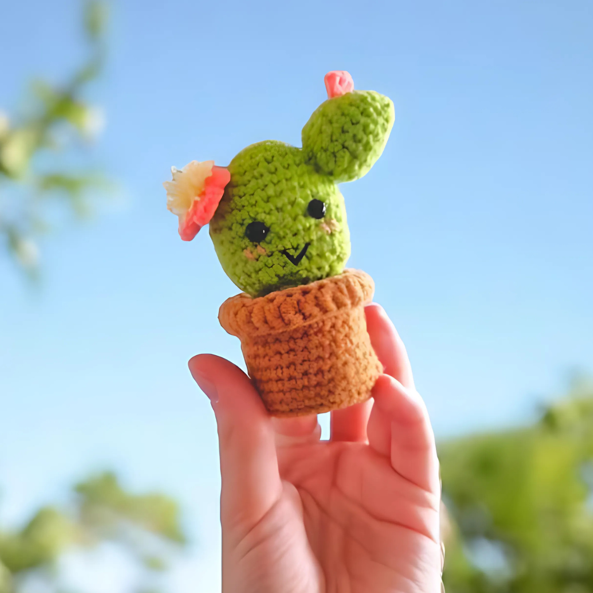 A hand holds up a smiling, flower-topped Crochet Cactus Kit 3pcs in a brown pot against a blue sky—an adorable crochet home decor piece.