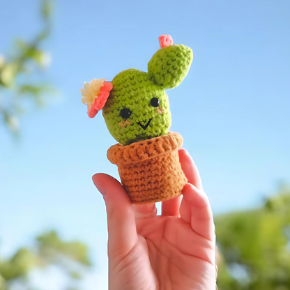 A hand holds up a smiling, flower-topped Crochet Cactus Kit 3pcs in a brown pot against a blue sky—an adorable crochet home decor piece.