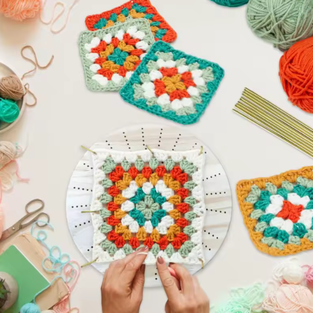 Hands working on a colorful granny square crochet piece surrounded by finished squares in a round grid layout, yarn balls, metal crochet hooks, and premium steel pins, all displayed on the Round Crochet Blocking Board.