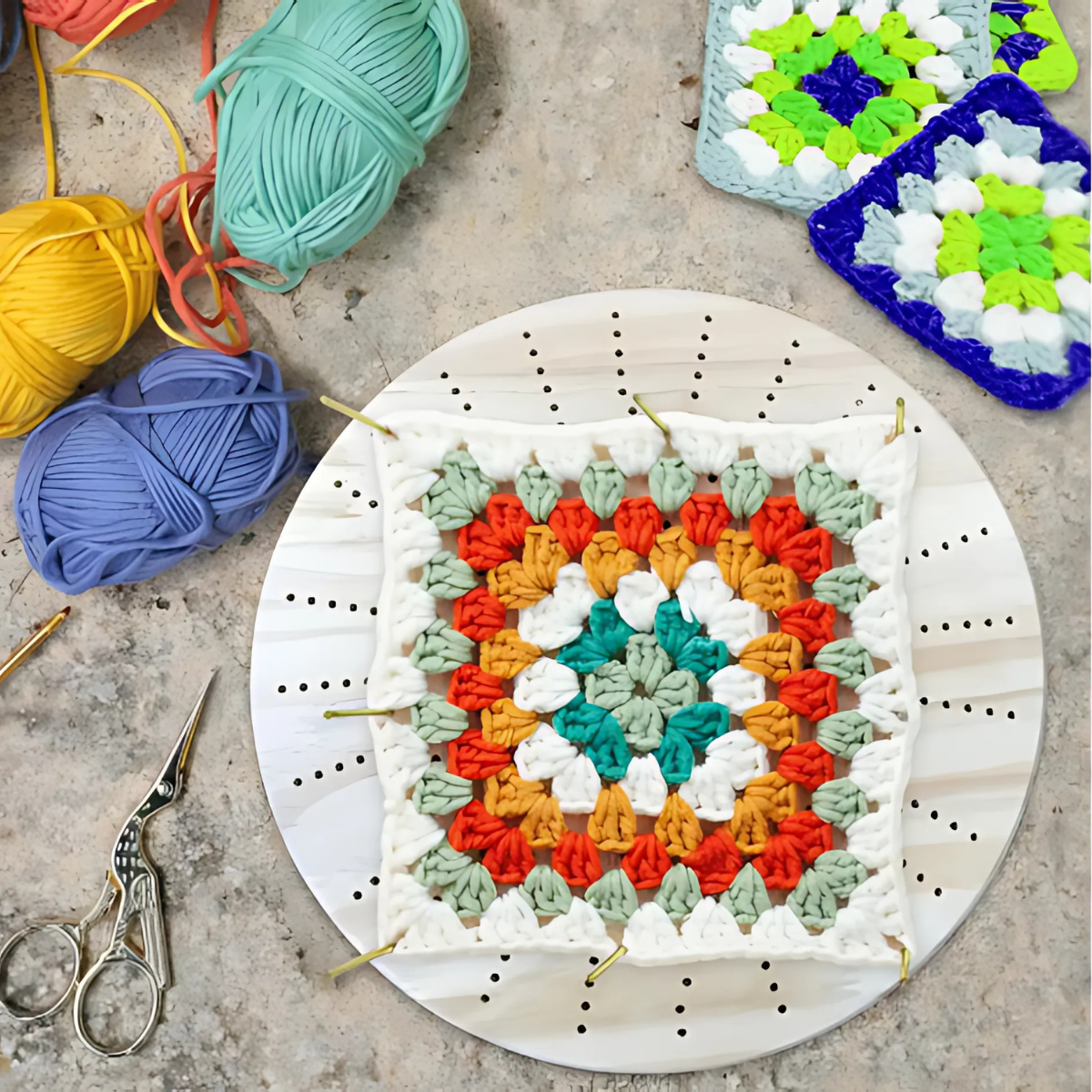 A crocheted granny square is pinned to the Round Crochet Blocking Board with a round grid, surrounded by colorful yarn balls, premium steel pins, scissors, and two extra granny squares.