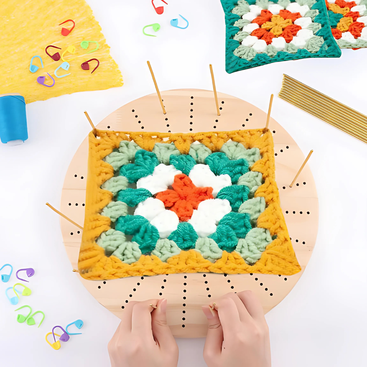 A granny square is pinned and stretched on the Round Crochet Blocking Board with a circular grid. Yarn, crochet tools, and stitch markers surround the board as two hands adjust it using premium steel pins.