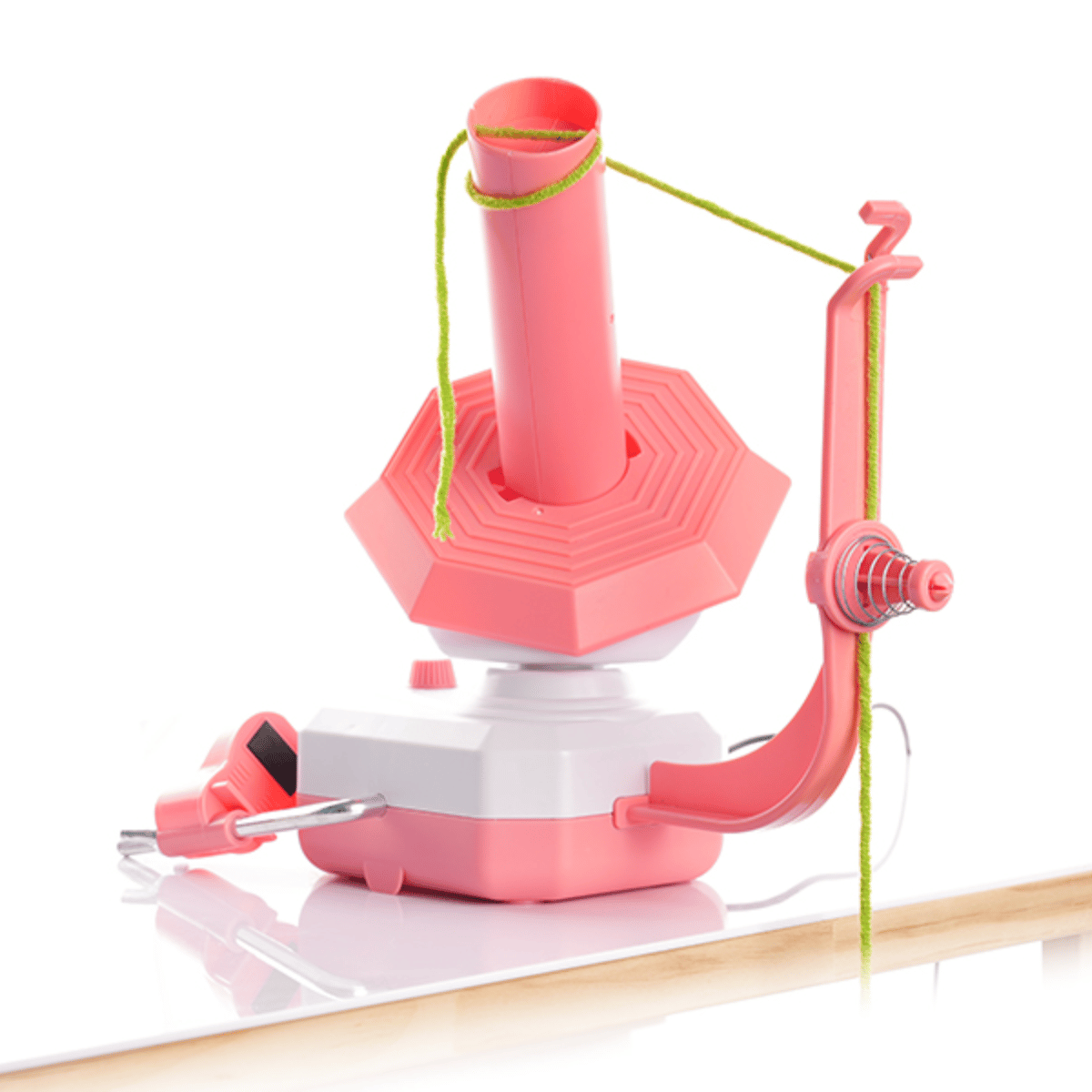 A pink and white Electric Yarn Winder with green yarn threaded through the guide, placed on a white reflective surface.