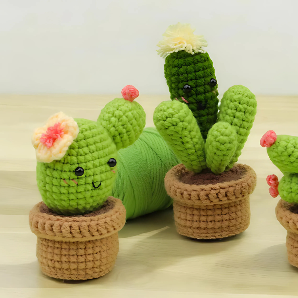 The Crochet Cactus Kit 3pcs features three smiling crocheted cactus toys with flowers in small brown pots, arranged on a light wooden surface with yarn in the background, perfect for charming home decor.