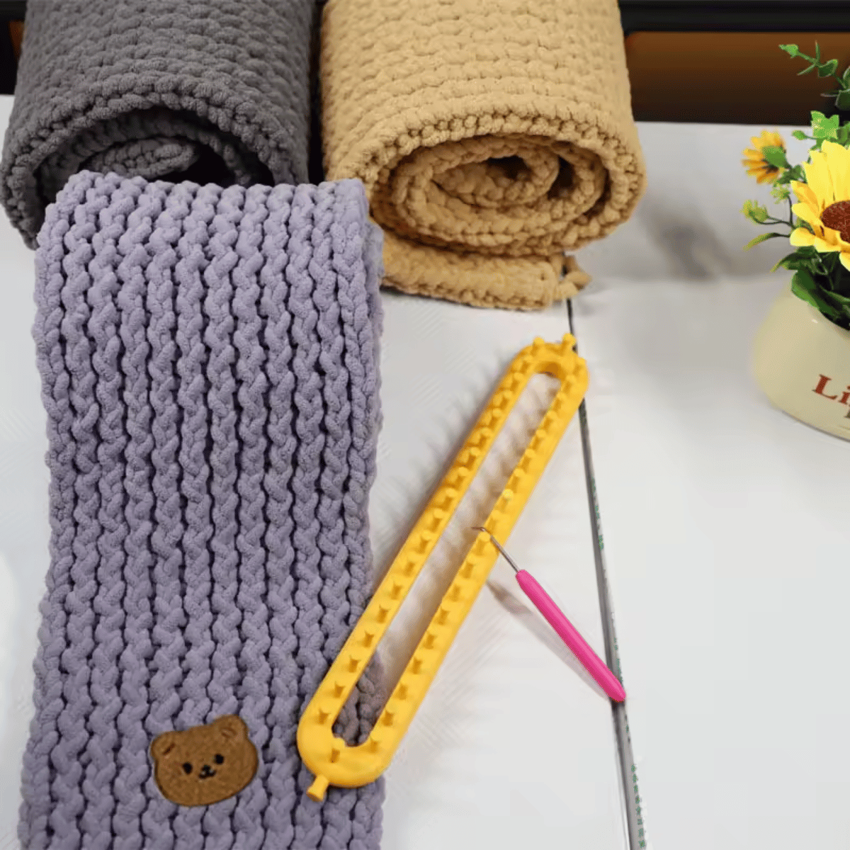 The Knitting Loom Set includes three chunky knit scarves in grey, beige, and lavender, a yellow beginner-friendly loom tool, a pink hook, and a pot of yellow flowers—perfect for quick and cozy knitting projects.