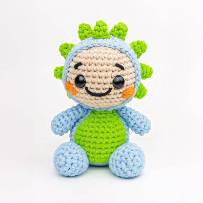 Spiky The Dino is a crocheted plush toy featuring a blue and green body, round black eyes, a smiling face, and spiky green details on its head, displayed against a plain white background.