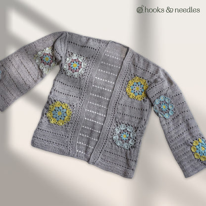 The Sunbloom Jacket Crochet Pattern features a gray crocheted cardigan made from organic cotton with yellow, blue, and white circular motifs on the sleeves and front, plus the "hooks & needles" logo on the top right.