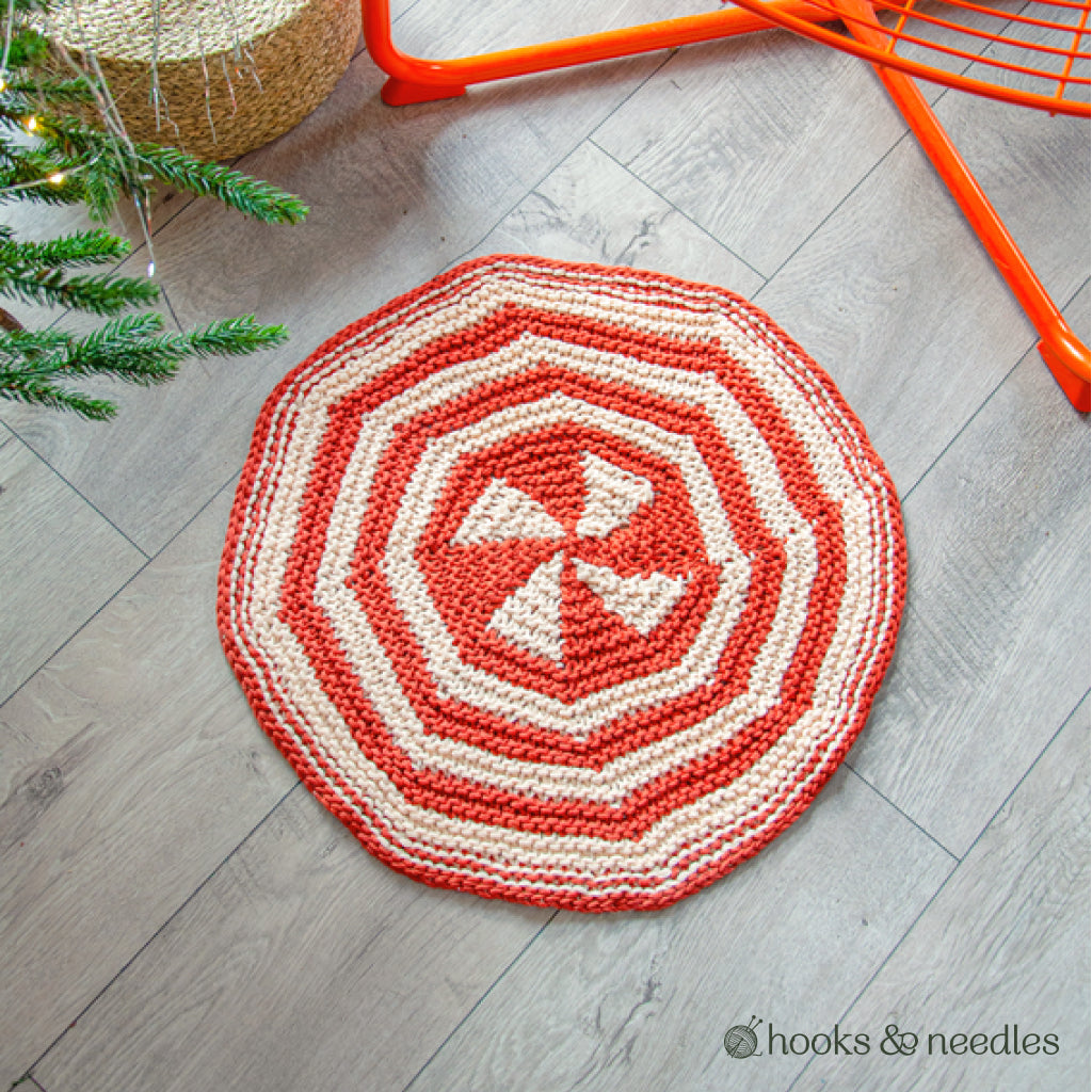 An Accent Rug Knitting Pattern with a round shape, red and white spiral and geometric design is displayed on a light wooden floor beside a Christmas tree and an orange chair.