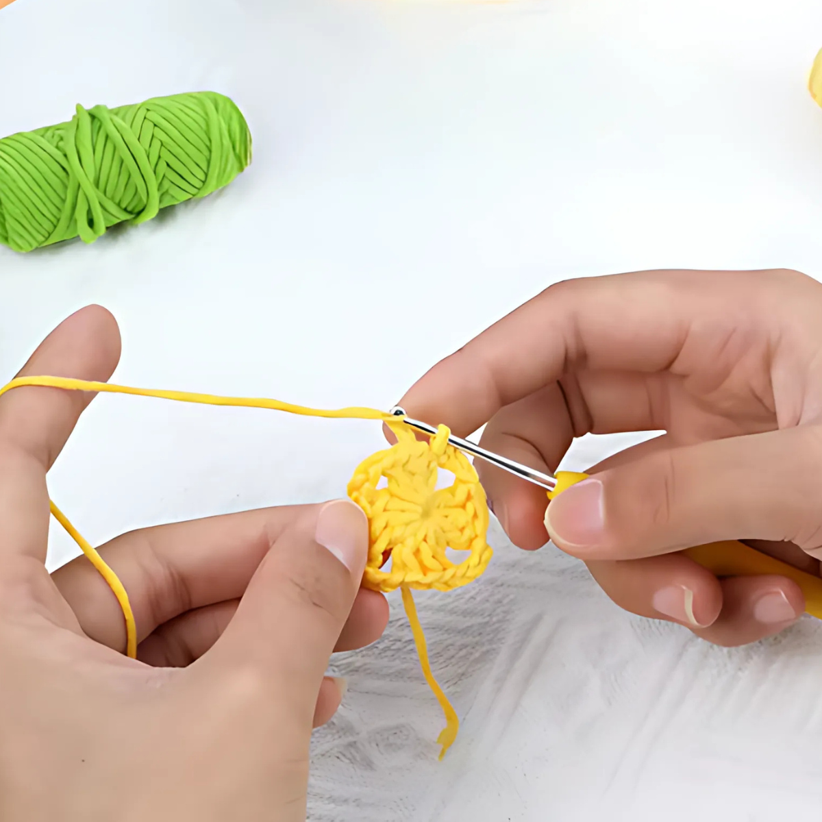 Two hands crochet a small yellow circle with a metal hook, while a green yarn ball sits nearby—ideal supplies included in the Crochet Flower Purse Kit.
