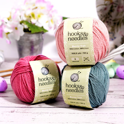 Three skeins of medium-weight, cotton-linen blend yarn in pink, red, and teal colors from the Limited Edition Easter Special Collection (bundle), displayed with knitting needles and soft-focus flowers in the background.