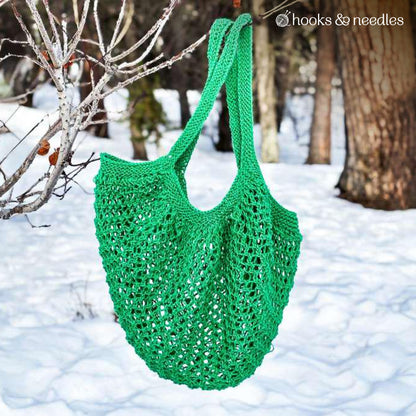 A hand-knitted Market Tote, crafted using the Market Tote Knitting Pattern, hangs from a tree branch in a snowy forest setting.