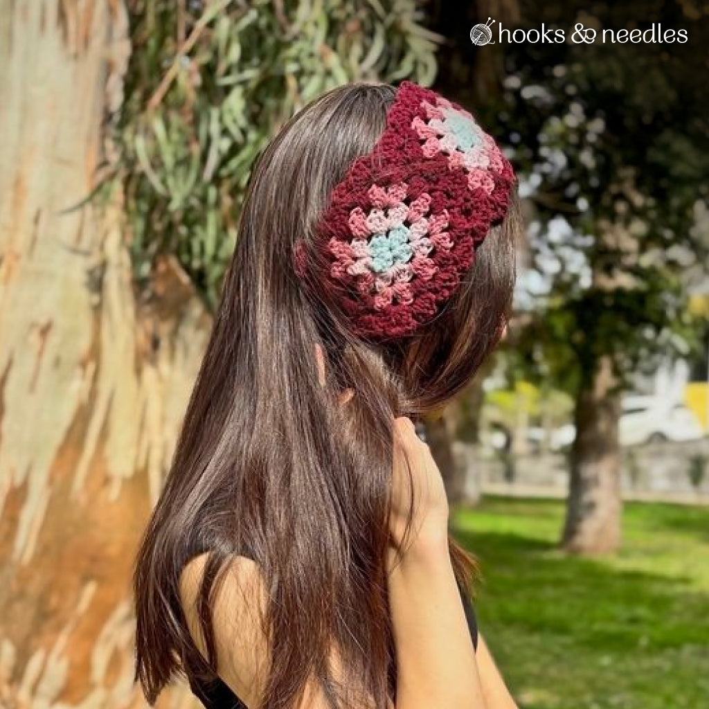 A person with long brown hair wears the Vintage Headband Crochet Pattern—a burgundy ear warmer with pink and green granny square accents—standing outdoors by a tree. The "hooks & needles" logo appears in the corner.