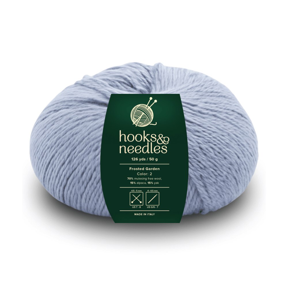 A skein of Frosted Garden in light blue, 126 yds/50g, features a green "hooks & needles" label and is crafted from ethical cotton and mulesing-free wool—ideal for mindful, cozy projects.
