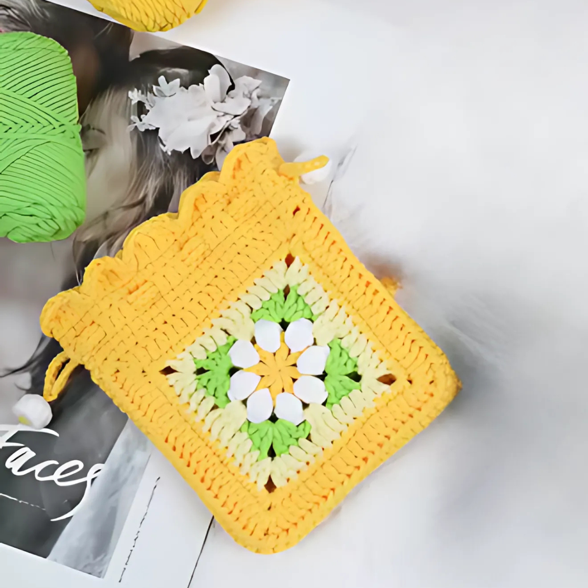 The Crochet Flower Purse Kit, featuring a yellow, green, and white floral square design, is displayed on a magazine beside a ball of green yarn.