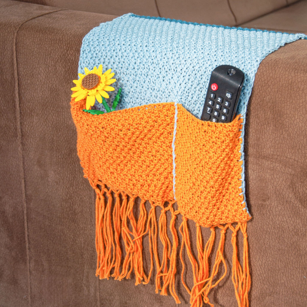 The Sofa Sidekick Knitting Pattern creates a vibrant organizer with orange pockets and fringe, perfect for draping over a brown sofa arm to hold essentials. Ideal for beginner knitters seeking style and function in one charming project.