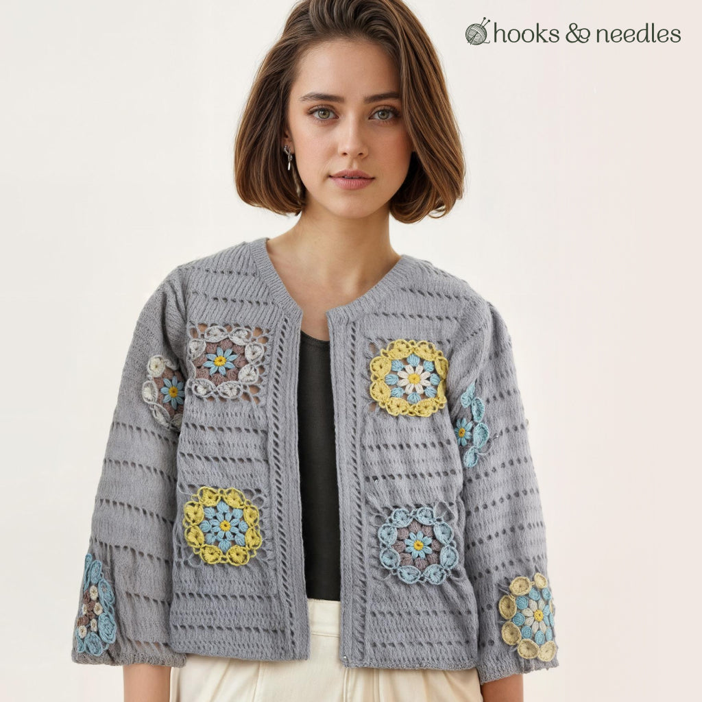 A woman models the Sunbloom Jacket Crochet Pattern in grey with colorful flower motifs, paired with a black top and white pants. "hooks & needles" appears in the top right against a plain background.