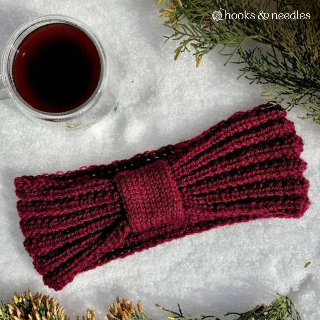 A burgundy rib stitch Woodland Whisper Ear Warmer Knitting Pattern is displayed on snow with a glass mug of tea, surrounded by pine branches.