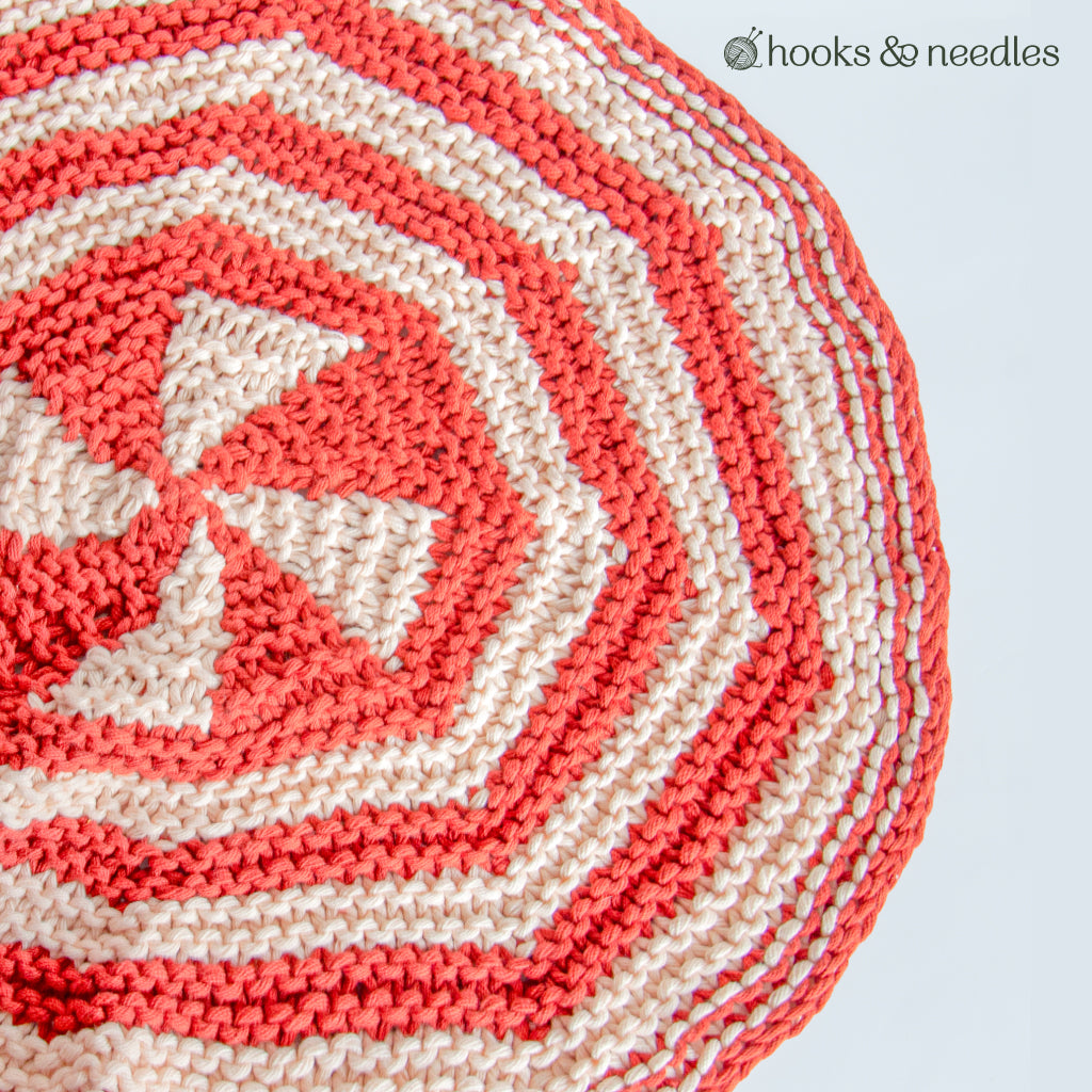 Close-up of the Accent Rug Knitting Pattern: a round, hand-knitted rug with spiral red and cream stripes, partially shown on a light background with "hooks & needles" in the corner.