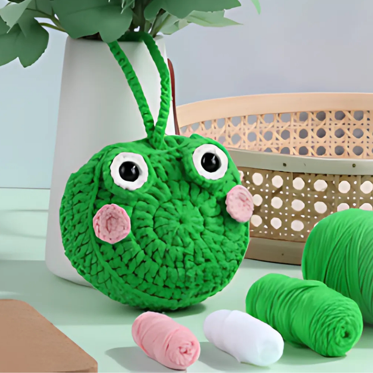 The Frog Storage Bag Crochet Kit features a green bag with pink cheeks and large eyes, displayed near yarn balls and a vase on a light green surface to showcase functional art.