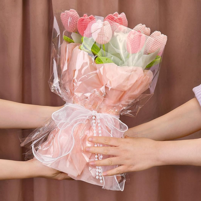 A hand presents a Crochet Tulip Bouquet Kit—pink and white crochet tulips wrapped in pink paper and clear plastic, tied with a white ribbon—to someone against a brown background.