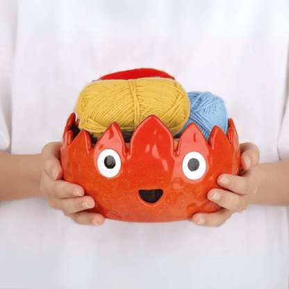 A person holds the Flame Ceramic Yarn Bowl, a red ceramic bowl with eyes and a mouth, containing yellow and blue yarn balls.
