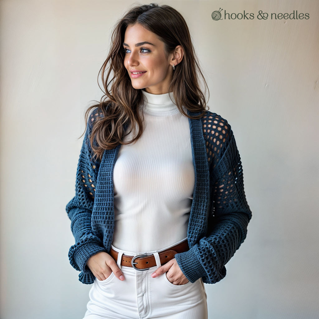 A woman in a white turtleneck and pants models the Honeycomb Stitch Cardigan Crochet Pattern—ideal for advanced crocheters. She stands indoors with hands in pockets. The Hooks & Needles logo is visible in the top right corner.