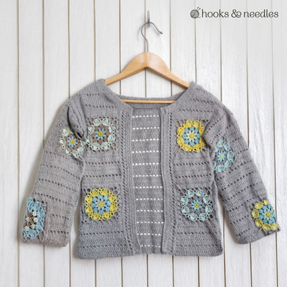 The Sunbloom Jacket Crochet Pattern is a gray organic cotton cardigan with vibrant granny square designs on the sleeves and front, displayed on a wooden hanger against a white wooden wall.