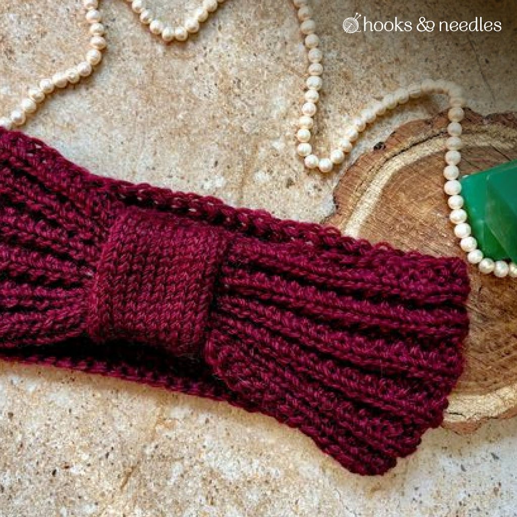 The Woodland Whisper Ear Warmer Knitting Pattern, featuring a maroon rib-stitched headband with a bow design, is shown on a tan surface beside a beaded necklace, wooden slice, green stone, and the "hooks & needles" logo.