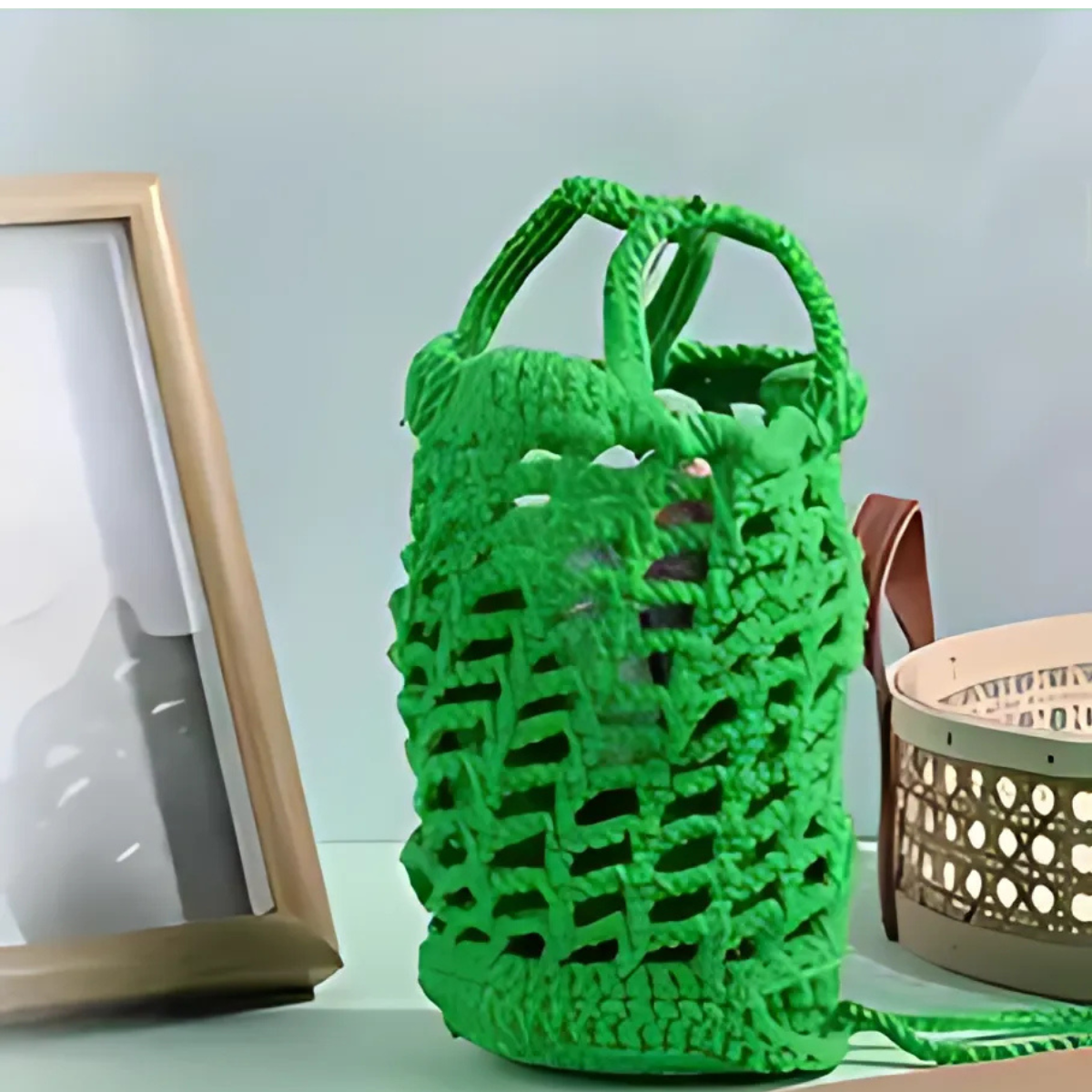 A Frog Storage Bag Crochet Kit sits on a surface beside a framed photo and a beige perforated container, blending functional art with playful style.