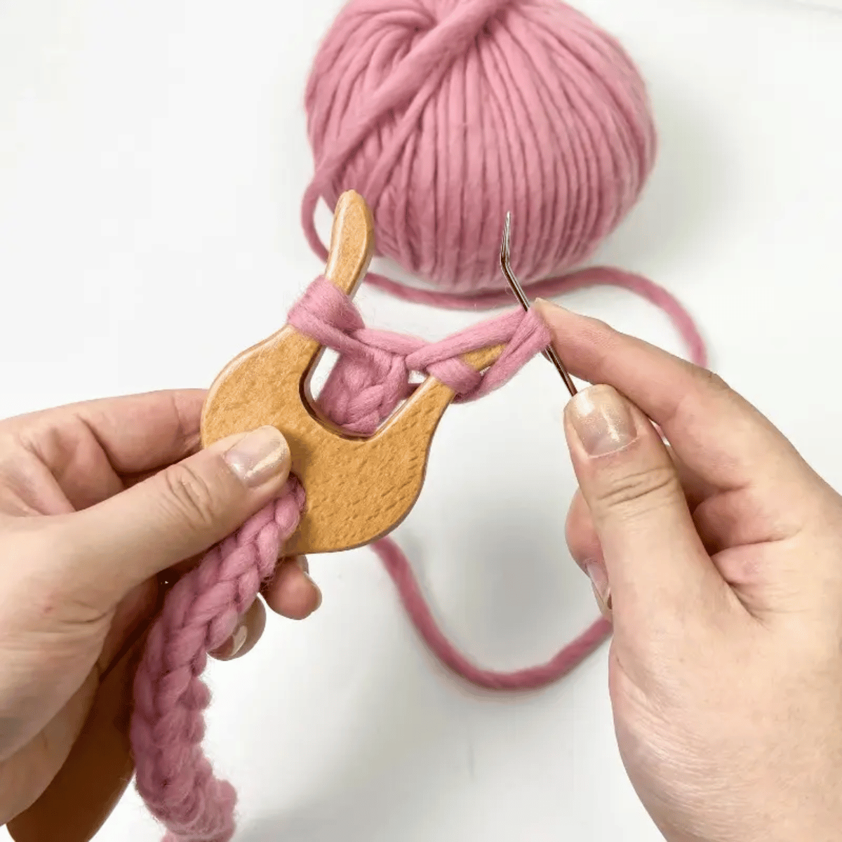 Hands using the Knitting Fork for Easy Cord Making with ergonomic design and hook to knit thick pink yarn, while a ball of yarn rests in the background on a white surface.