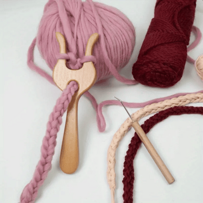 The Knitting Fork for Easy Cord Making, featuring an ergonomic wooden design, is shown with pink yarn, surrounded by skeins of yarn and a crochet hook on a white surface.