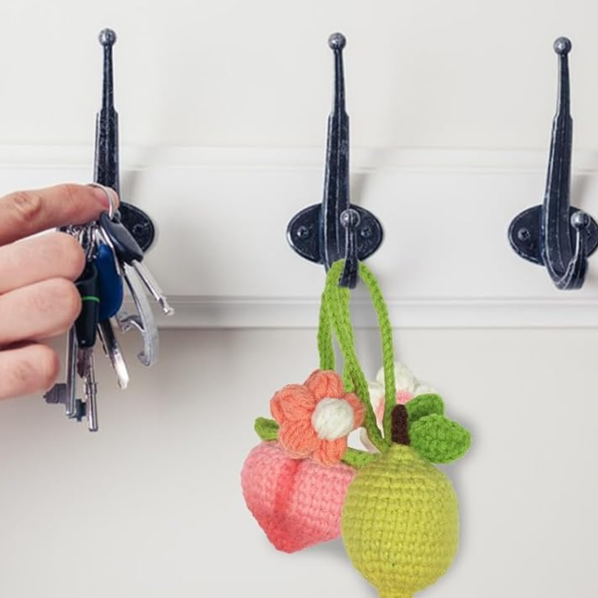 A hand hangs keys on a wall hook next to two crochet fruit pendants—a peach with flower and a lemon with leaves—made using the Crochet Fruits Kit 4pcs DIY set.
