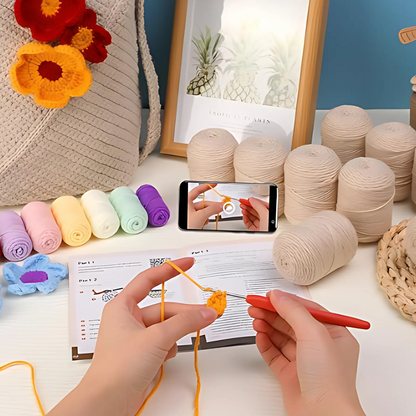 Crocheting with yellow yarn from the Bohemian Crochet Tote Bag Kit while watching a video tutorial on a smartphone. Yarn balls, crocheted flowers, and the kit’s pattern sheet are on the table.
