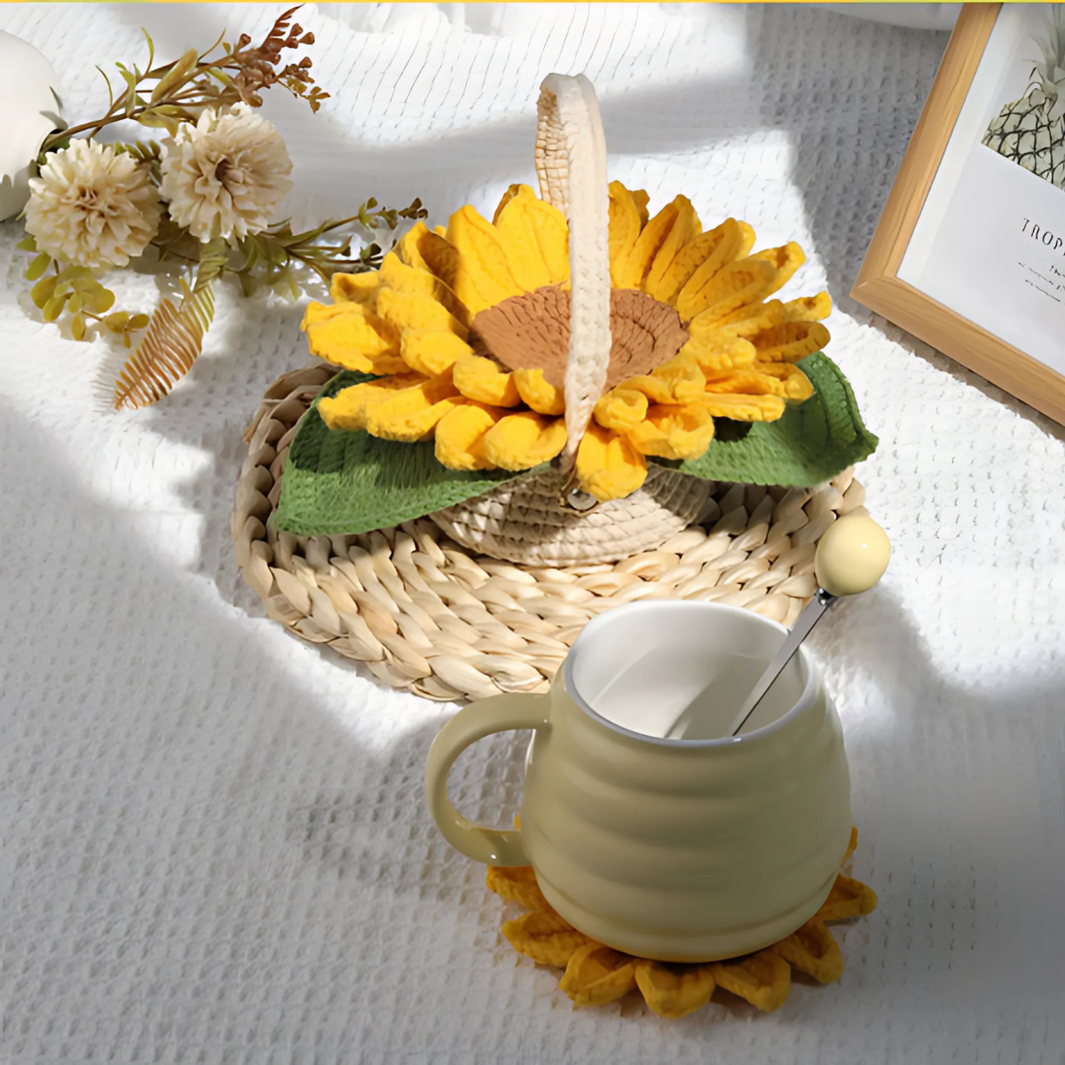 A yellow mug with a spoon rests on a handmade coaster from the Sunflower Pot Crochet Kit with 5pcs Coasters, placed on a white textured surface next to a sunflower basket, artificial flowers, and a framed picture.