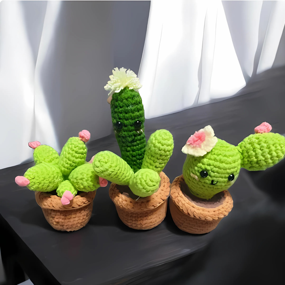 The Crochet Cactus Kit 3pcs features three unique crocheted cacti in small brown pots with decorative flowers and embroidered faces—charming crochet decor for any space. White fabric drapes in the background, enhancing their adorable appeal.
